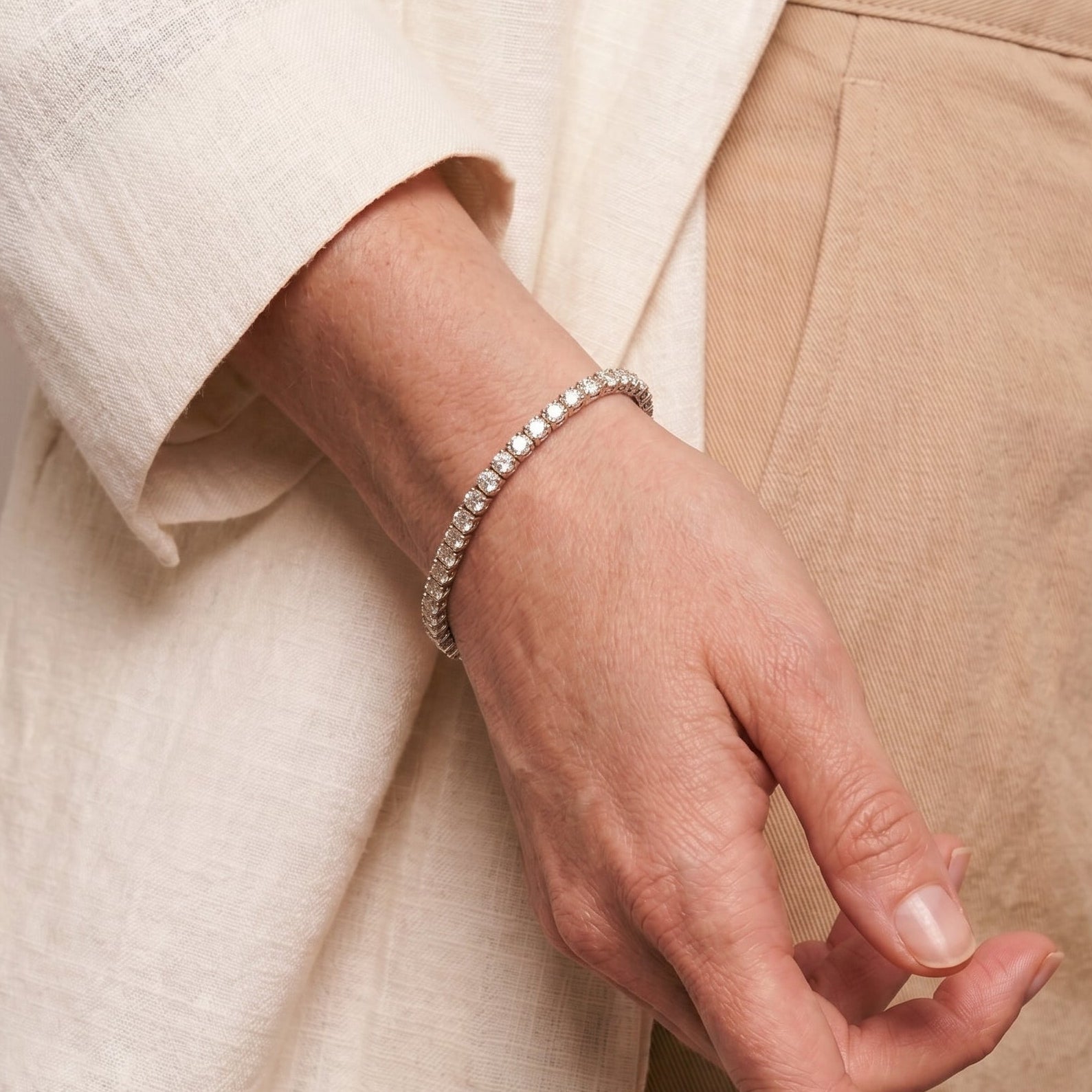 A close-up of a person's hand wearing a delicate silver tennis bracelet, styled with a cream linen top and beige trousers.