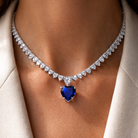 Necklace with a blue heart-shaped gemstone and diamond-studded chain on a person wearing a white coat.